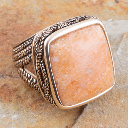 A square bezel-set ring featuring a warm peach-orange stone, set in a textured silver-toned metal band with a polished gold-toned bezel, resting on a textured stone surface.