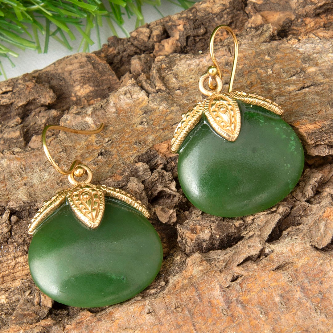 A pair of gold and green drop earrings displayed on a textured piece of wood. Each earring features a smooth, oval-shaped dark green stone pendant with a textured gold leaf-like cap and a gold fishhook ear wire.