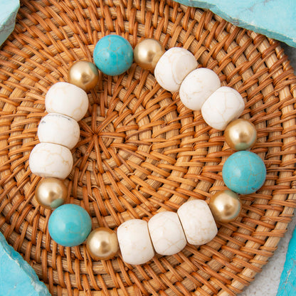 A beaded bracelet arranged in a circle on a woven wicker tray, featuring matte turquoise-blue beads, white marbled beads, and metallic gold beads.