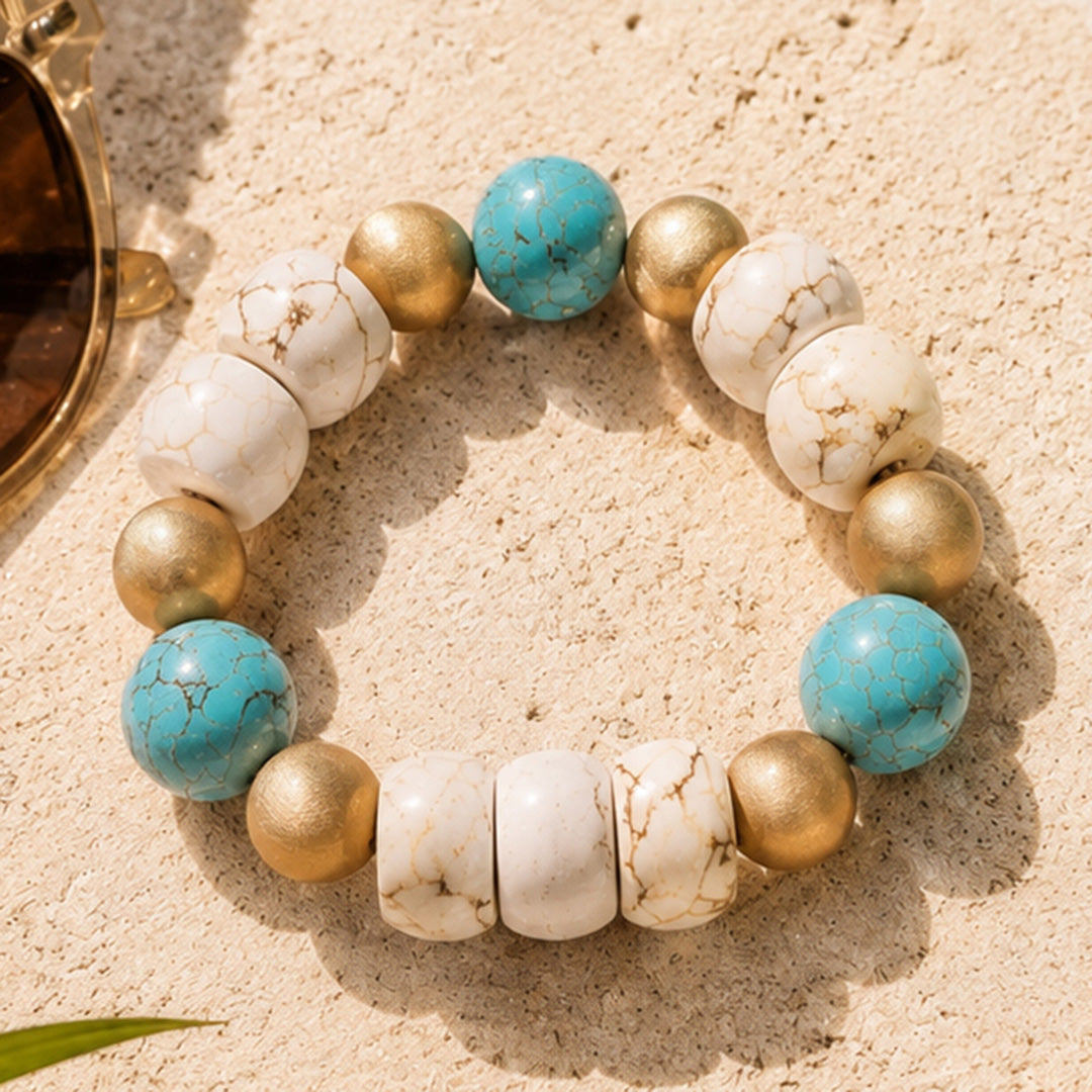 A beaded stretch bracelet featuring turquoise, white marble, and metallic gold beads, arranged in a circle on a sandy beige background.
