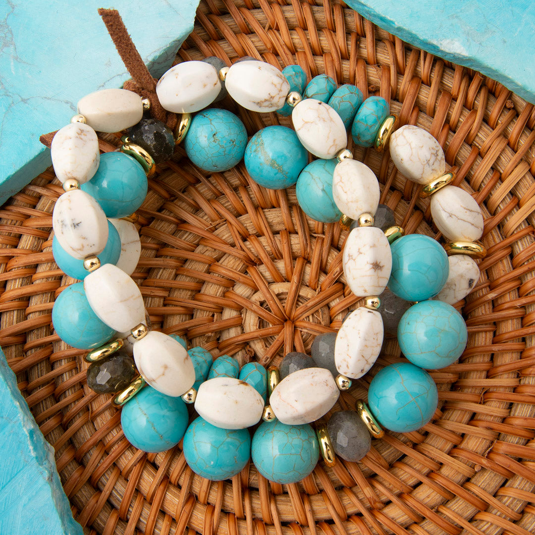 A beaded stretch bracelet featuring light blue turquoise, cream-colored marbled stone beads, and small gray faceted beads separated by gold-tone metal spacers, displayed on a woven wicker mat.