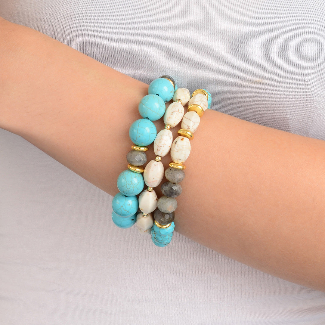 Three beaded stretch bracelets stacked on a person's wrist. The bracelets feature light blue, white, gray, and gold-toned beads.
