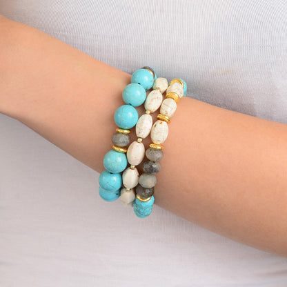 Three beaded stretch bracelets stacked on a person's wrist. The bracelets feature light blue, white, gray, and gold-toned beads.
