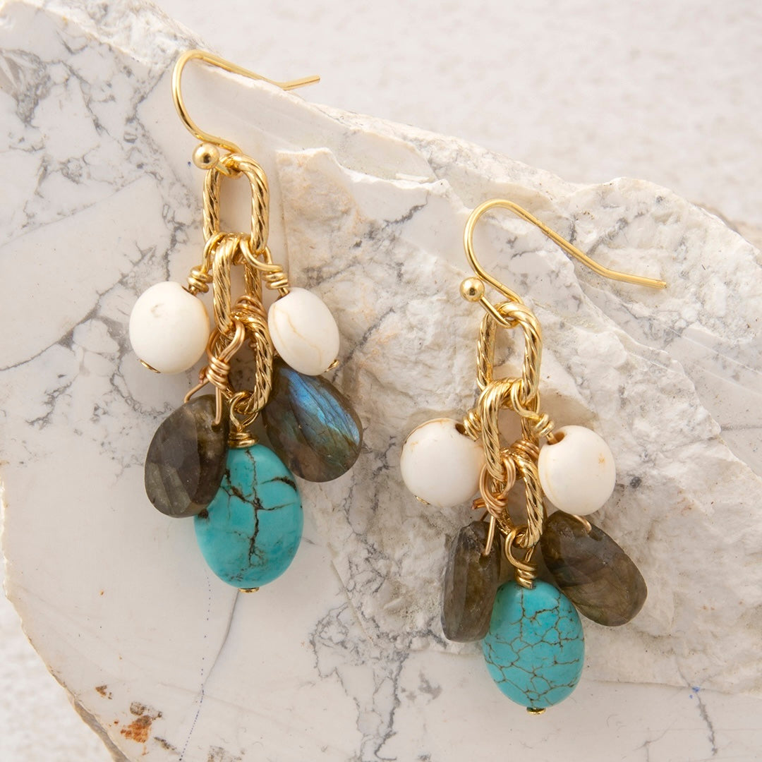 A pair of handmade dangle earrings with gold-tone wire wrapping, featuring white beads, dark gray labradorite beads, and turquoise and labradorite drop beads, displayed on a white marble slab.