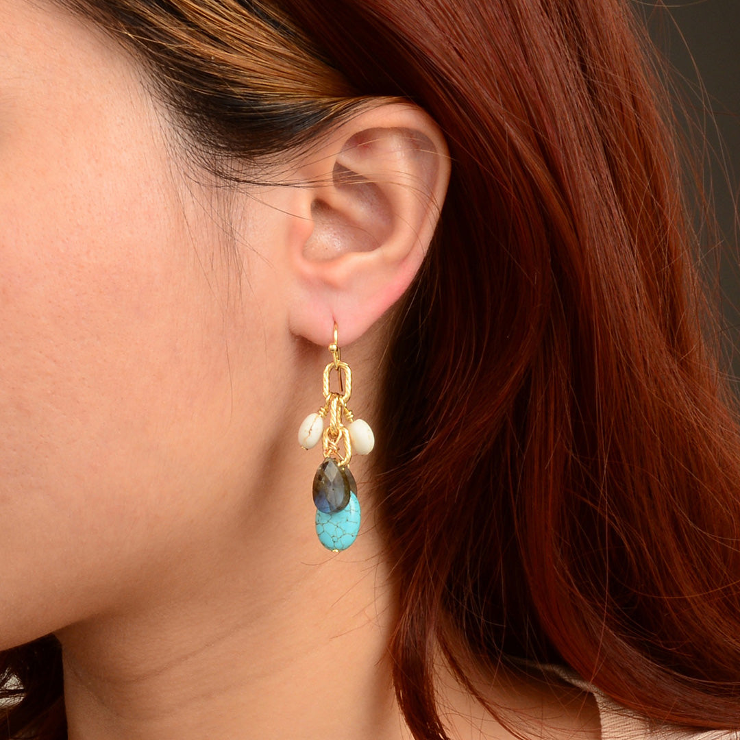 A close-up of a person's ear wearing a gold dangle earring with white beads, a dark gray stone, and a turquoise blue stone.