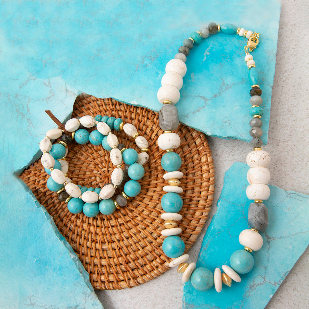 A bohemian-style beaded jewelry set including a necklace and bracelet, featuring light blue, white, gray, and gold beads, styled on a woven wicker mat over a textured light blue background.