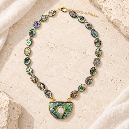 A gold-tone necklace with irregular abalone shell beads and a large semicircle pendant, styled on a textured beige stone surface with draped white fabric.