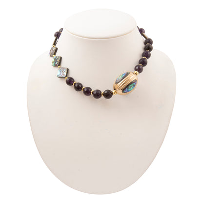 A beaded necklace displayed on a white jewelry bust. The necklace features dark, nearly black round beads, several iridescent shell beads with blue and green tones, and a prominent gold-toned oval bead with blue-green inlay.