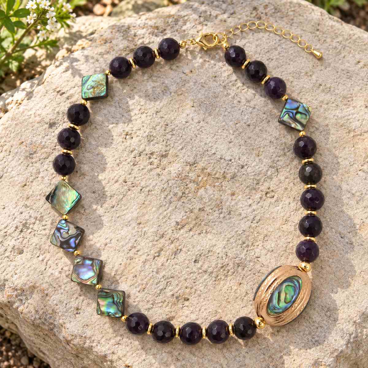 A beaded necklace with dark purple faceted beads, iridescent green and blue abalone shell square tiles, and a large oval abalone shell centerpiece, displayed on a light beige rock outdoors.