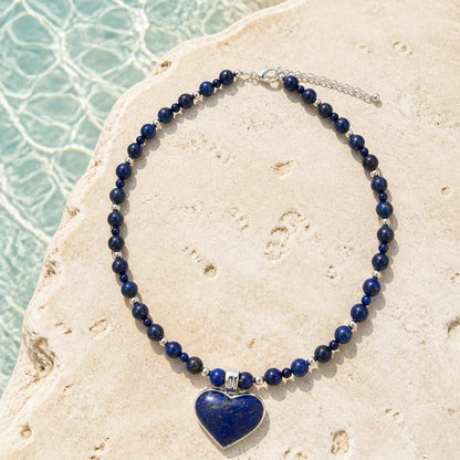 A beaded necklace with dark blue lapis lazuli stones and a heart-shaped pendant, displayed on a light beige stone surface with water in the background.