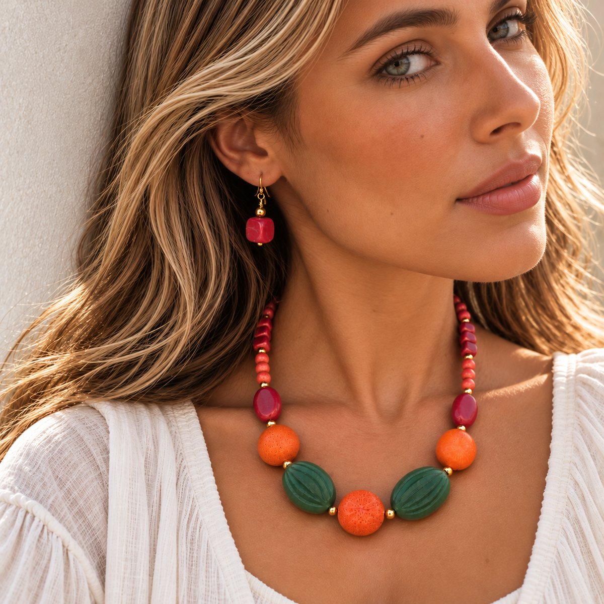 A woman wearing a colorful statement necklace and matching drop earrings. The necklace features green leaf-shaped beads, orange round beads, and magenta beads with gold-tone connectors.