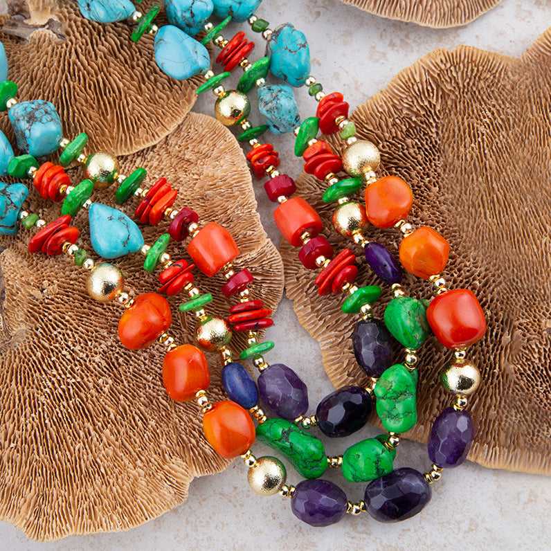 A close-up of a multi-strand beaded necklace laid on brown textured mushroom caps over a light stone surface. The necklace features turquoise, coral, green, purple, orange, and gold beads.